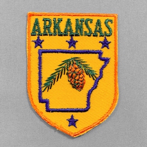 May include: Embroidered patch featuring the word "ARKANSAS" in green above a yellow shield. The shield has a blue outline of the state with a pinecone and leaves. Three blue stars are above the state outline, and one star is below.