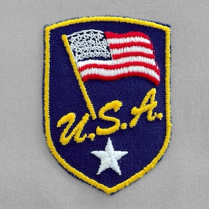 May include: A shield-shaped embroidered patch with a dark blue background and a yellow border. It features a waving American flag, the yellow embroidered text U.S.A., and a single white star centered at the bottom of the patch.
