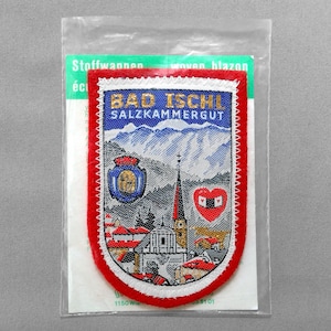 May include: A woven patch featuring the text "BAD ISCHL SALZKAMMERGUT" above a mountain scene and a church. The patch has a red border and is packaged in a clear plastic bag.