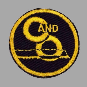 May include: Embroidered patch with a black background and a yellow border. The patch features the letters "C" and "O" in yellow, with the word "AND" in between. A yellow line and shape are at the bottom.