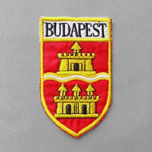 May include: Embroidered patch featuring the word "BUDAPEST" above a red shield design. The shield displays two yellow castle illustrations with black details, set against a red background, and outlined in yellow.