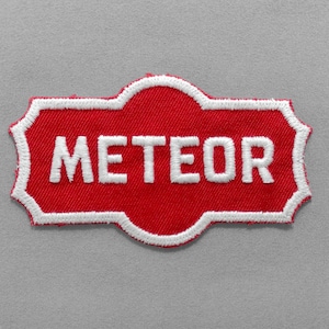 May include: A red embroidered patch with the word "METEOR" in white block letters. The patch has a white border and a unique, decorative shape. The background is a light gray fabric.