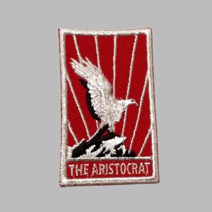 May include: Embroidered patch featuring a white eagle with black accents perched on a mountain, set against a red background with white rays. The text "THE ARISTOCRAT" is displayed at the bottom, framed by a white border.