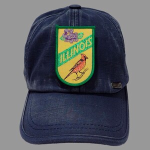 May include: A blue denim baseball cap with a yellow and green patch featuring a cardinal bird, a purple flower, and the text "ILLINOIS".