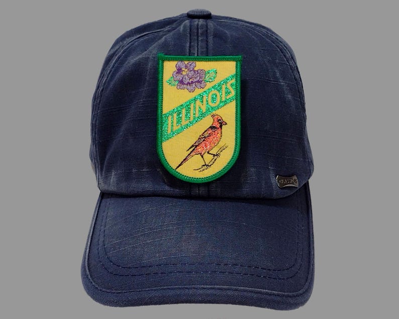 May include: A blue denim baseball cap with a yellow and green patch featuring a cardinal bird, a purple flower, and the text "ILLINOIS".