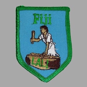 May include: Embroidered patch with a shield shape, featuring a light blue background and a green border. The patch displays the word "Fiji" and an illustration of a person working with a wooden object. The word "LALI" is also present.
