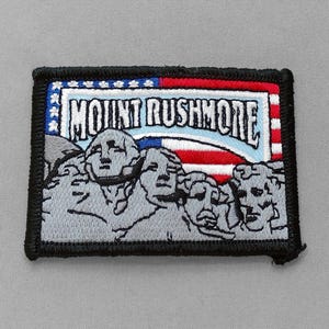 May include: Embroidered patch featuring Mount Rushmore with the text "MOUNT RUSHMORE". The patch has a black border and incorporates the American flag's colors: red, white, and blue. The image depicts the faces of the presidents.