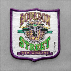 May include: A shield-shaped embroidered patch with a white background and a purple border. It features the text BOURBON STREET NEW ORLEANS in purple and green. A central graphic depicts a stylized mask with yellow and purple striped patterns.