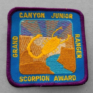 May include: A blue and purple embroidered patch with the text "Grand Canyon Junior Ranger Scorpion Award". The patch features a yellow scorpion silhouette against a colorful background.