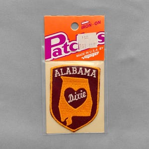 May include: An iron-on embroidered patch in a shield shape with a maroon background and a gold outline. The patch features a gold map of Alabama with a central heart containing the word Dixie in white script, and the word ALABAMA in white text.