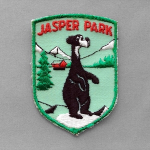 May include: A shield-shaped embroidered patch featuring a black bear standing on white snow against a light green background. The text JASPER PARK is stitched in red at the top. The design includes a small red cabin, green pine trees, and white mountains.