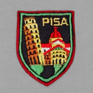 May include: A red, green, and black embroidered patch with the word "PISA" in white lettering. The patch features a depiction of the Leaning Tower of Pisa and a church.