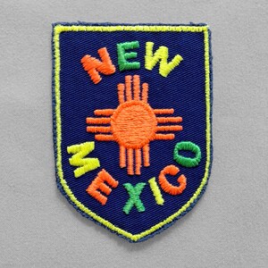 May include: Embroidered patch featuring the state of New Mexico. The patch is a shield shape with a navy blue background. The words "NEW MEXICO" are embroidered in orange, green, and yellow. The state symbol is in the center.