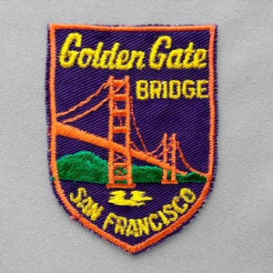 May include: A shield-shaped embroidered patch featuring a purple background with an orange border. It depicts the Golden Gate Bridge in orange, green hills, and yellow water. The text reads Golden Gate BRIDGE and SAN FRANCISCO in yellow.