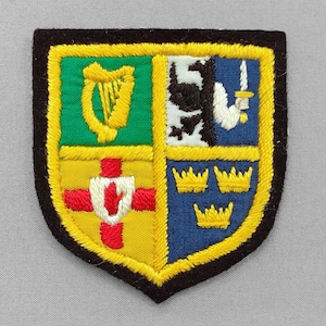 May include: Embroidered patch with a shield design. The shield is divided into four sections, featuring a gold harp on green, a black and white cow, a white arm holding a sword on blue, and three gold crowns on blue. The patch has a black border with yellow stitching.