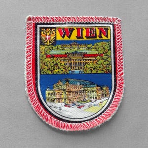 May include: A rectangular embroidered patch with a red stitched border. The design features the text WIEN in red letters, a golden double-headed eagle emblem, and two detailed illustrations of historic buildings in Vienna, Austria.