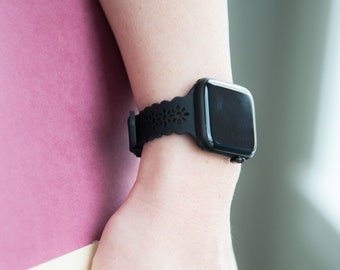 Könnte beinhalten: Eine schwarze Smartwatch mit quadratischem Zifferblatt und dekorativem schwarzen Armband ist zu sehen. Die Uhr befindet sich an einem Handgelenk, und die Person hält ein rosa-cremefarbenes Buch.