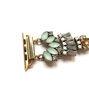 Puede incluir: Una pulsera de reloj dorada con un cierre decorativo con diamantes de imitaci&oacute;n verde claro y transparentes.