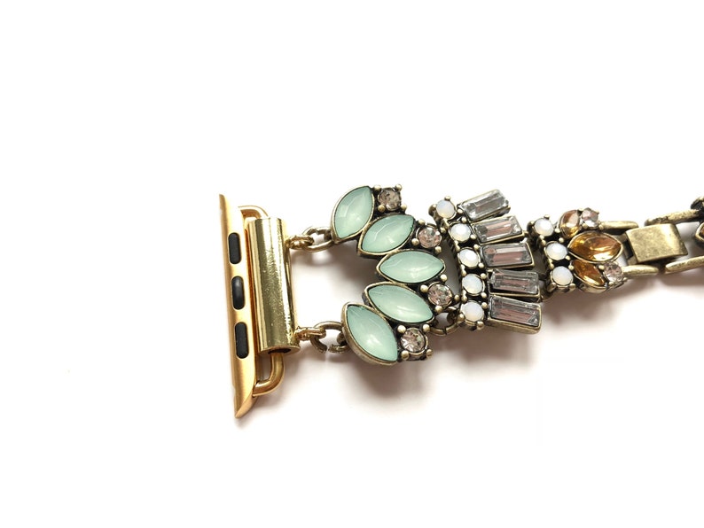 Puede incluir: Una pulsera de reloj dorada con un cierre decorativo con diamantes de imitaci&oacute;n verde claro y transparentes.