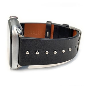 May include: A silver Apple Watch with a black leather band featuring silver studs. The band has a brown leather lining.