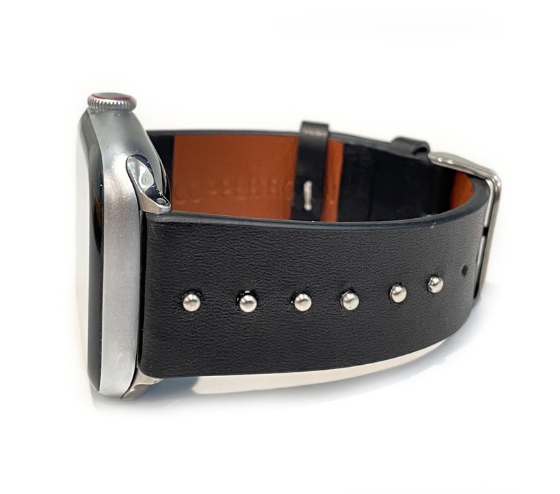 May include: A silver Apple Watch with a black leather band featuring silver studs. The band has a brown leather lining.