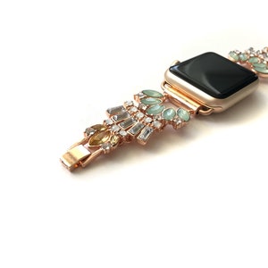 Puede incluir: Una banda de reloj Apple de oro rosa con una esfera negra y un dise&ntilde;o decorativo con diamantes de imitaci&oacute;n transparentes, verdes y amarillos.