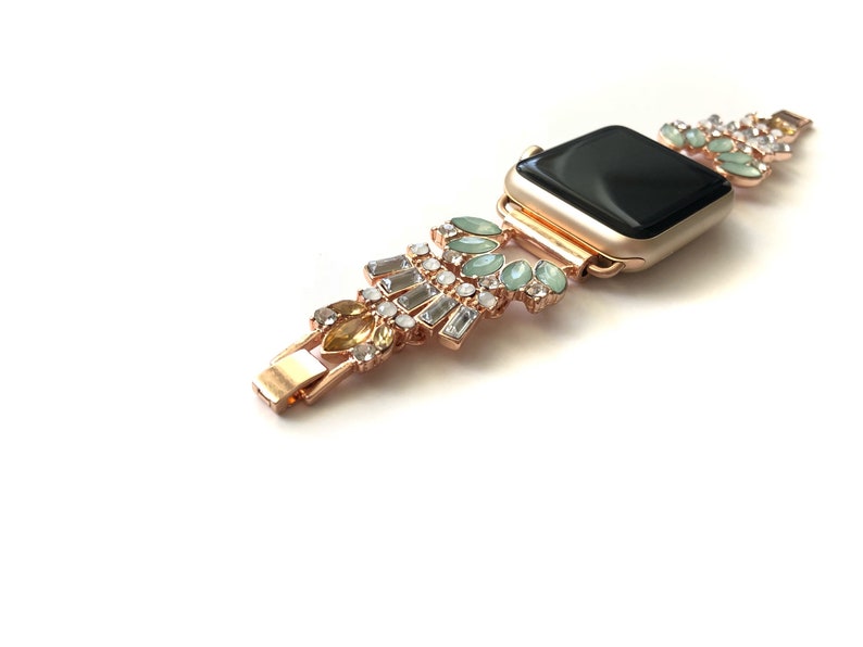 Puede incluir: Una banda de reloj Apple de oro rosa con una esfera negra y un dise&ntilde;o decorativo con diamantes de imitaci&oacute;n transparentes, verdes y amarillos.