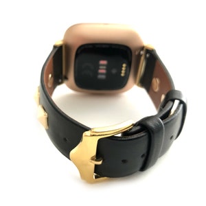 Może przedstawiać: Złoty zegarek smartwatch z czarnym sk&oacute;rzanym paskiem i złotymi akcentami. Pasek ma złote ćwieki i złotą klamrę.