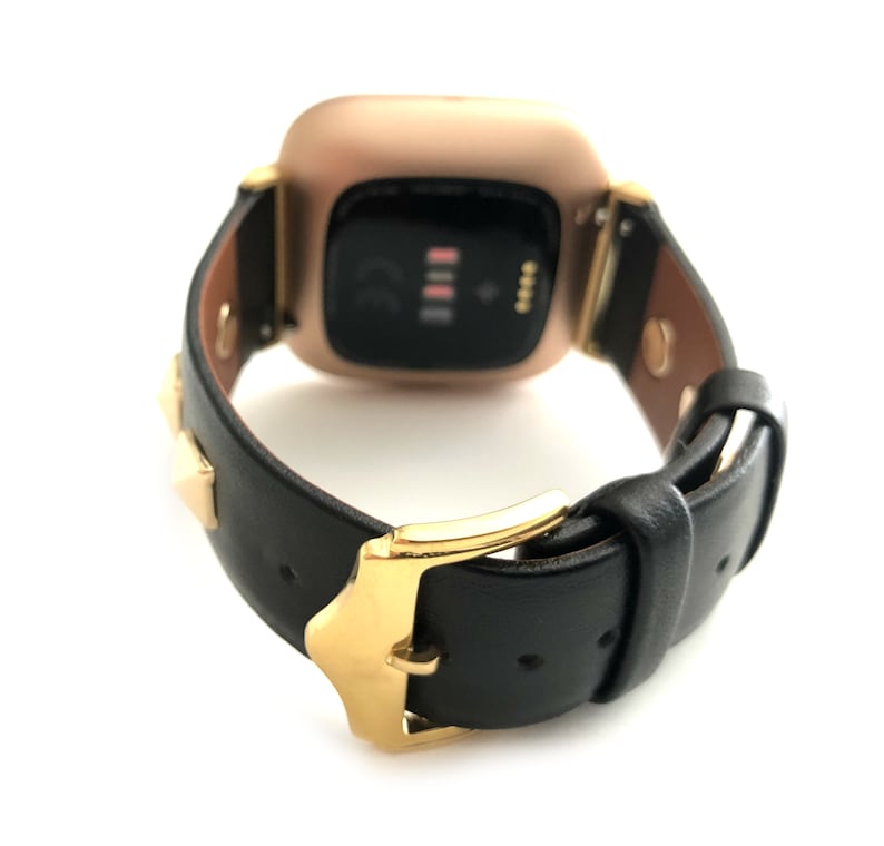Może przedstawiać: Złoty zegarek smartwatch z czarnym sk&oacute;rzanym paskiem i złotymi akcentami. Pasek ma złote ćwieki i złotą klamrę.