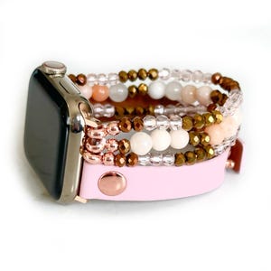 May include: A smartwatch band featuring a light pink leather strap with a rose gold metal snap button. The band is attached to a metallic watch face and includes multiple strands of beads in clear, opaque white, soft peach, and metallic bronze tones.
