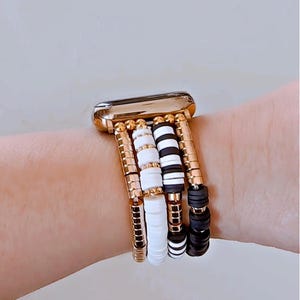 May include: A gold Apple Watch with a white and black beaded band. The band is made of multiple strands of beads, including white round beads, black round beads, and black and white striped beads.