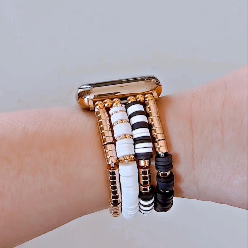 May include: A gold Apple Watch with a white and black beaded band. The band is made of multiple strands of beads, including white round beads, black round beads, and black and white striped beads.