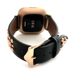 May include: A black leather watch band with rose gold accents and studs. The band is designed for a smartwatch with a square face.