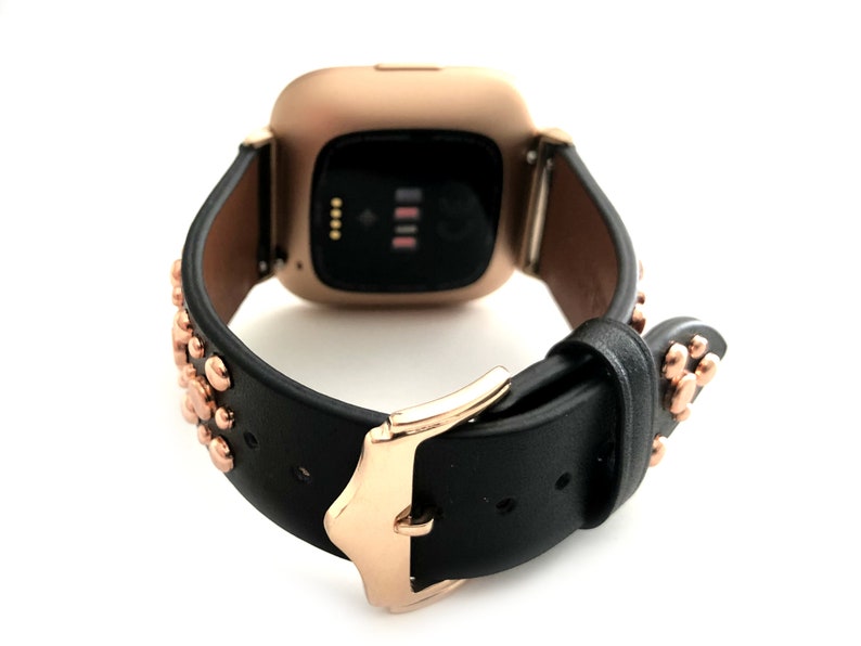 May include: A black leather watch band with rose gold accents and studs. The band is designed for a smartwatch with a square face.