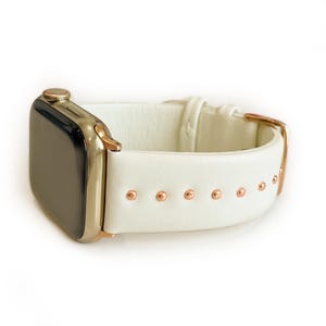 May include: White leather Apple Watch band with rose gold accents and studs.