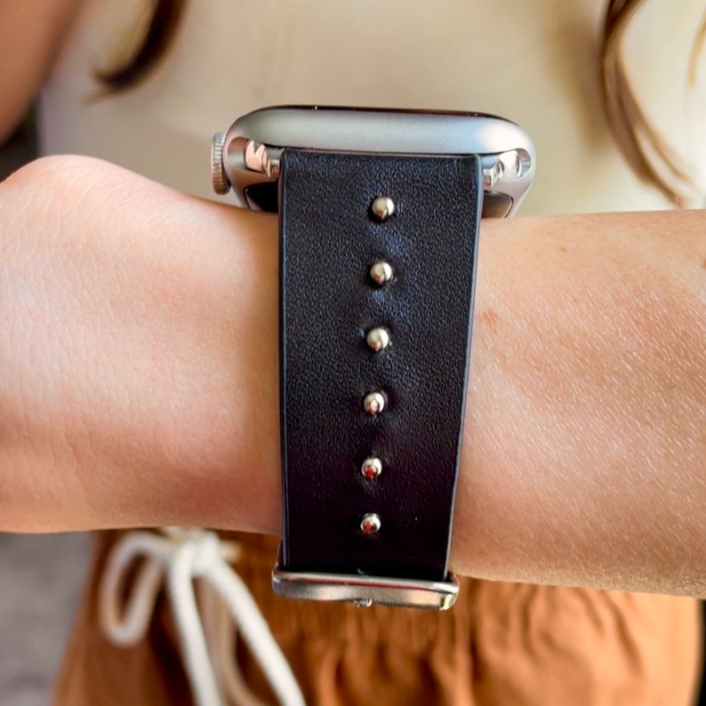 May include: A silver Apple Watch with a black leather band featuring silver studs.