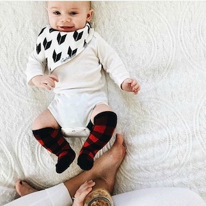 May include: A baby wearing a white long-sleeved onesie with a black and white patterned bib. The baby is wearing red and black plaid leg warmers. The baby is lying on a white blanket.