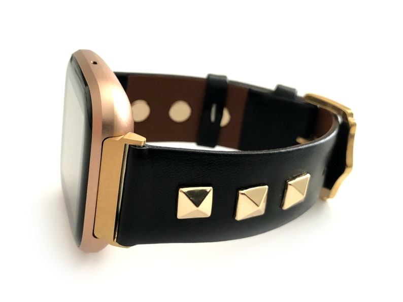 May include: A rose gold smartwatch with a black leather band featuring gold pyramid-shaped studs.