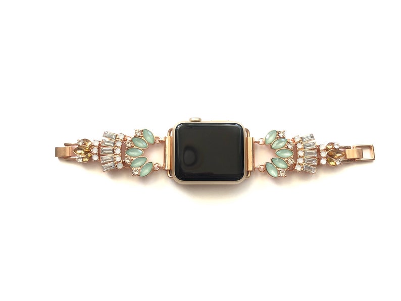Puede incluir: Una banda de reloj Apple de oro rosa con una esfera negra. La banda est&aacute; adornada con diamantes de imitaci&oacute;n transparentes y verdes.