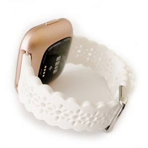 May include: A white fitness tracker band with a rose gold watch face. The band has a delicate floral pattern.