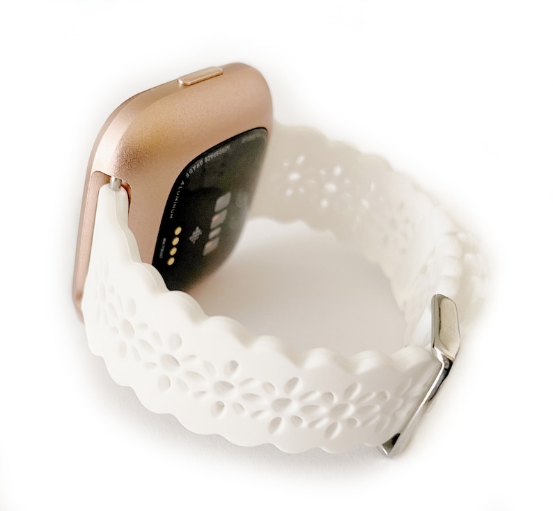 May include: A white fitness tracker band with a rose gold watch face. The band has a delicate floral pattern.