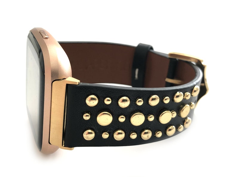 May include: A black leather watch band with gold studs. The band is designed for a square-shaped smartwatch.