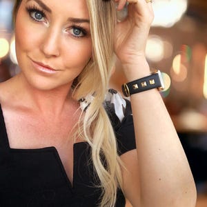May include: A woman wearing a black v-neck top with a black and gold studded watch on her wrist.