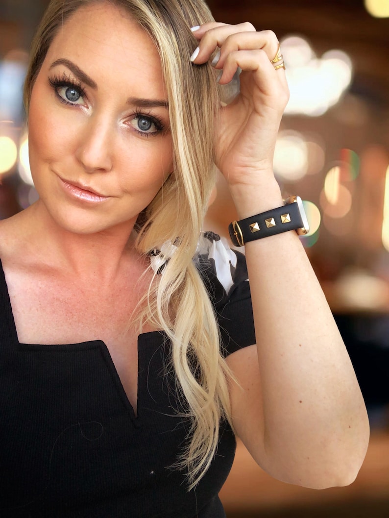 May include: A woman wearing a black v-neck top with a black and gold studded watch on her wrist.