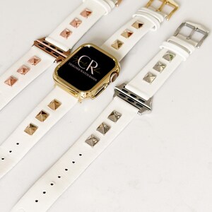 May include: Three white leather Apple Watch bands with gold and silver pyramid-shaped studs. The bands are shown on a white surface. The middle band has a gold Apple Watch face with the text "CR Designer Watch Bands".