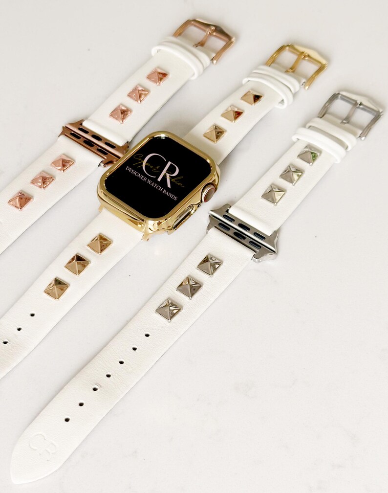 May include: Three white leather Apple Watch bands with gold and silver pyramid-shaped studs. The bands are shown on a white surface. The middle band has a gold Apple Watch face with the text "CR Designer Watch Bands".