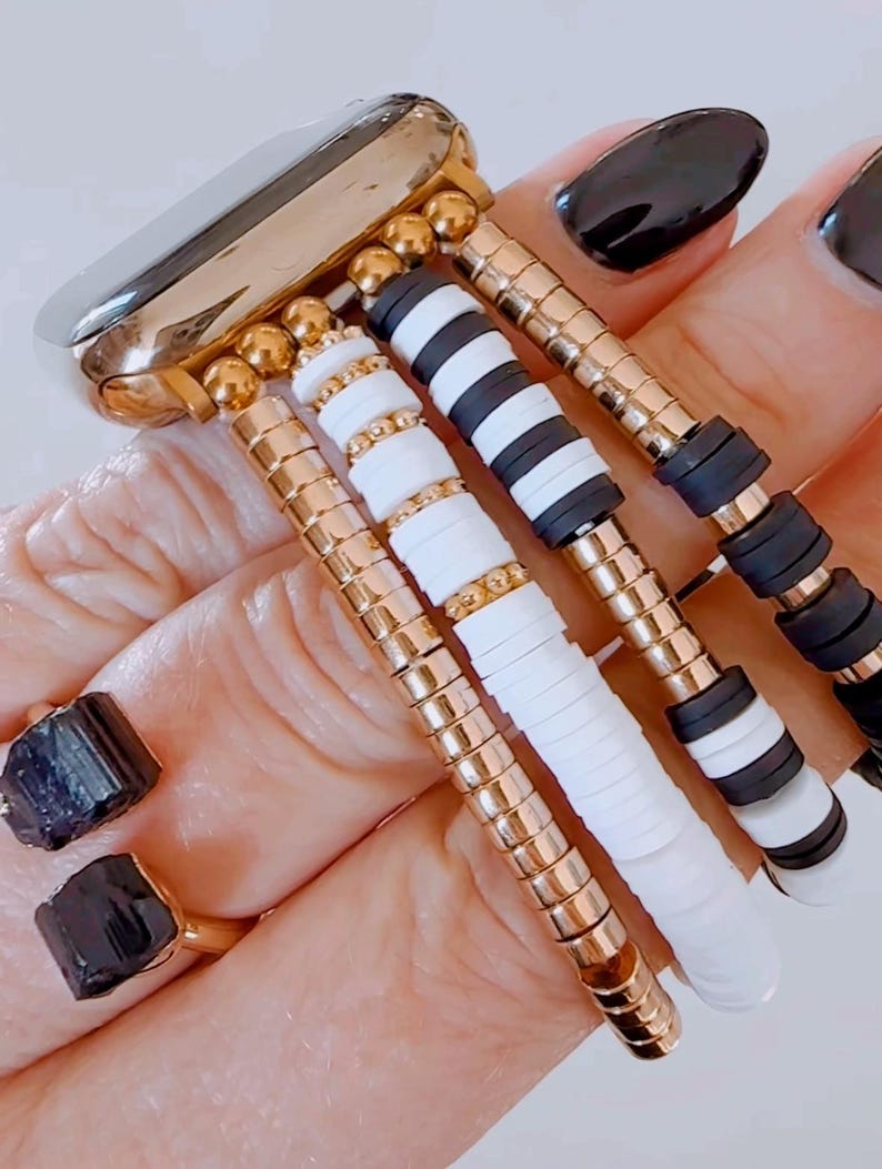 May include: A gold watch band with multiple black and white beaded bracelets attached. The bracelets are made of small round beads and flat, rectangular beads.