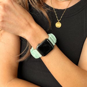 Puede incluir: Una correa de reloj tipo scrunchie verde menta combinada con un reloj inteligente negro. Tambi&eacute;n se ve un collar de cadena dorada con un colgante redondo. La correa del reloj est&aacute; hecha de una tela suave y fruncida.