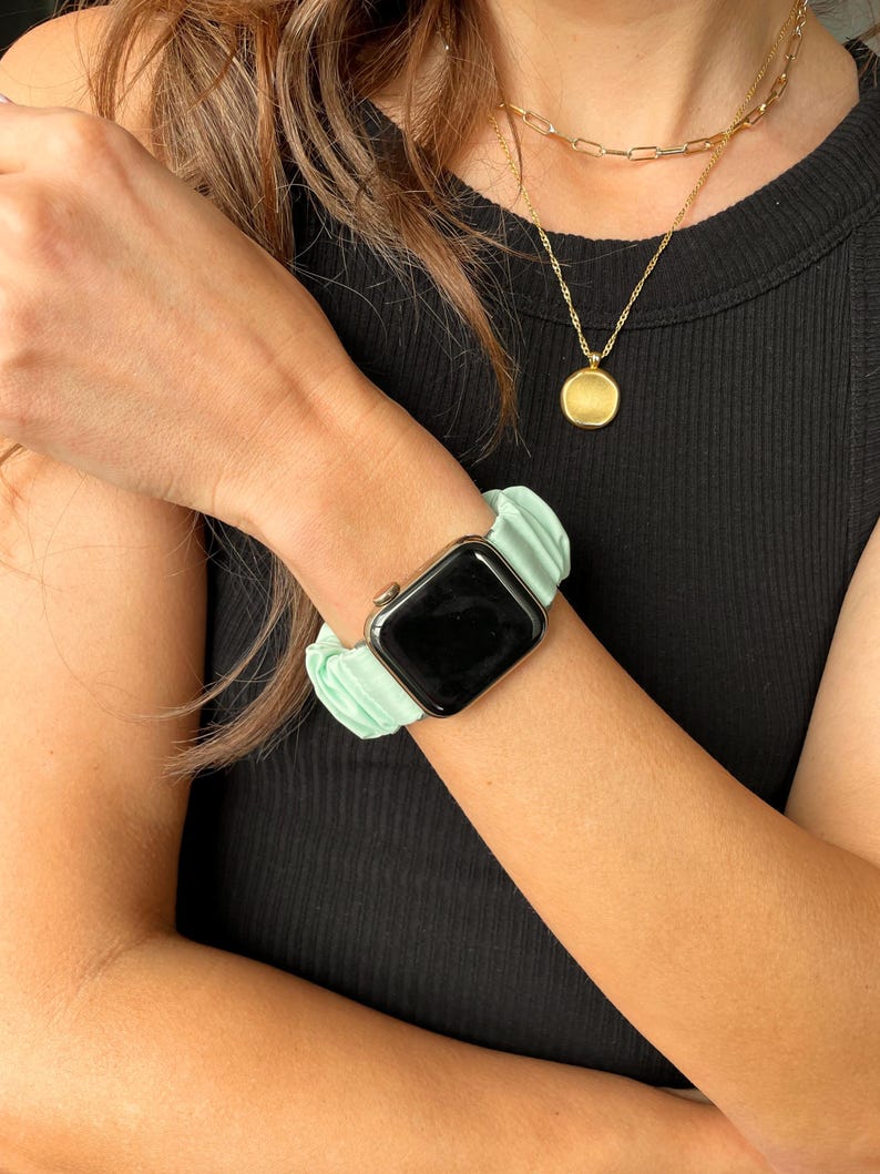 Puede incluir: Una correa de reloj tipo scrunchie verde menta combinada con un reloj inteligente negro. Tambi&eacute;n se ve un collar de cadena dorada con un colgante redondo. La correa del reloj est&aacute; hecha de una tela suave y fruncida.