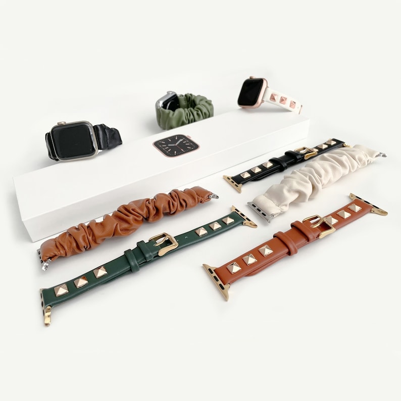 Op de afbeelding: Een assortiment Apple Watch-bandjes in diverse kleuren en stijlen. De afbeelding toont bandjes in bruin, groen, zwart en cr&egrave;mekleurig leer, versierd met goudkleurige studs. Scrunchie-stijl bandjes en een zwarte leren band zijn ook te zien.